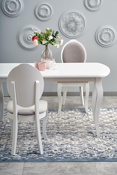 Na zdjęciu jest prezentowany asortyment sklepu oficjalnego producenta stołów i krzeseł DDMeble. Jest na nim wnętrze w stylu glamour z szarą farbą i okrągłymi dekoracjami zdobionymi ornamentami kwiatowymi na ścianach. Z białym stołem o zaokrąglonym blacie na szczytach i nogami w typie cabriole. Z białymi krzesłami z owalnym oparciem i półowalnym siedziskiem tapicerowane beżowym materiałem. Stoją na ozdobnym dywanie o mozaikowej teksturze.  